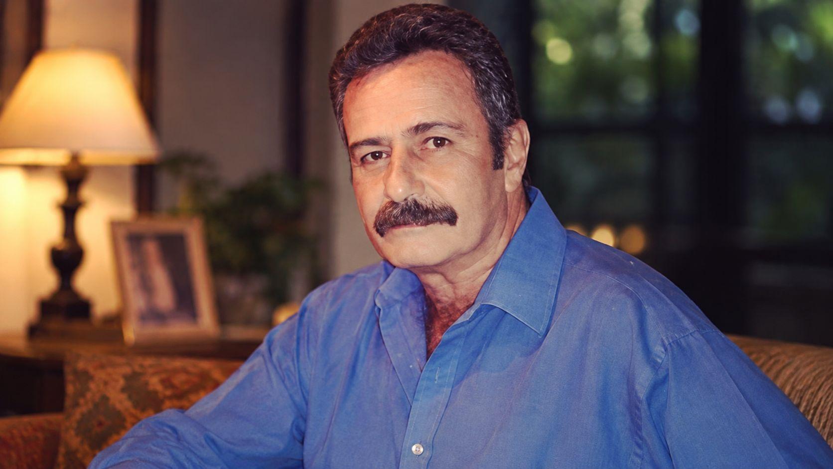 Retrato del actor mexicano Carlos Miguel con camisa azul y bigote, sentado en un ambiente interior con iluminación cálida.